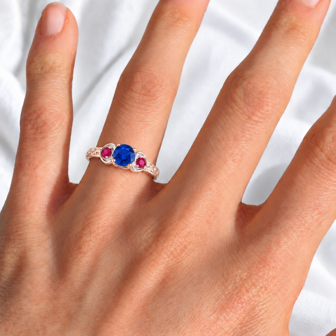 Sapphire and ruby three-stone ring with leaf band in 14K rose gold, worn on hand